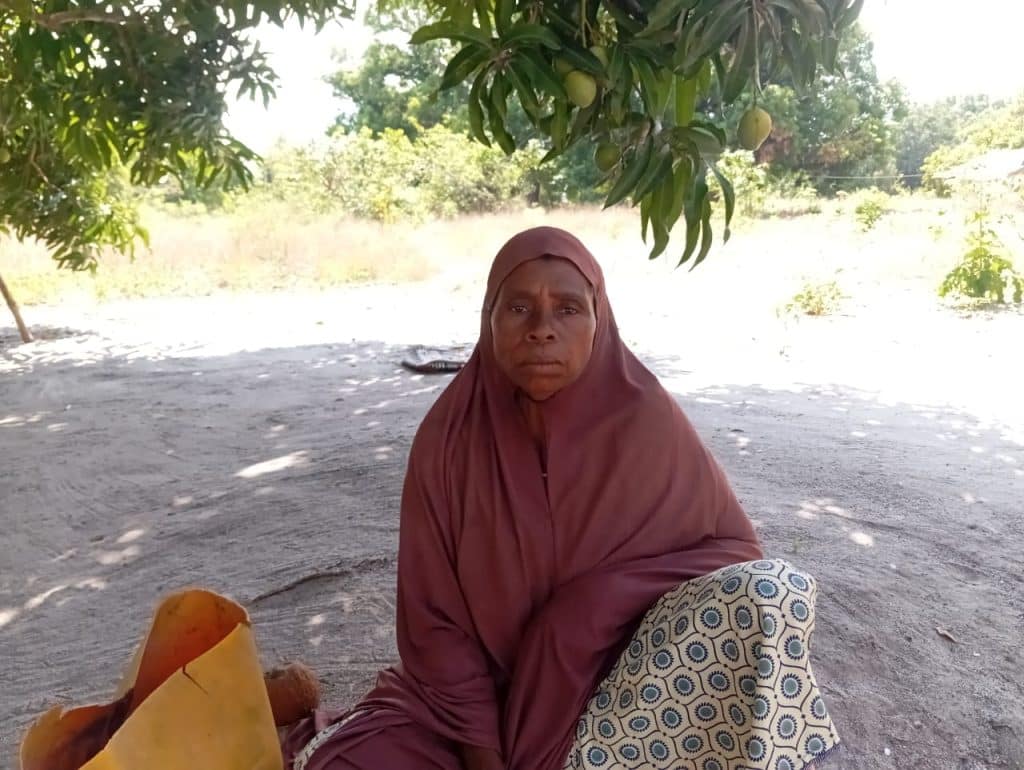Middle-aged woman sitting on the ground under a tree in a rural area, wearing a headscarf and patterned skirt, with a serious expression, representing poverty, unemployment, and resilience in an African community.