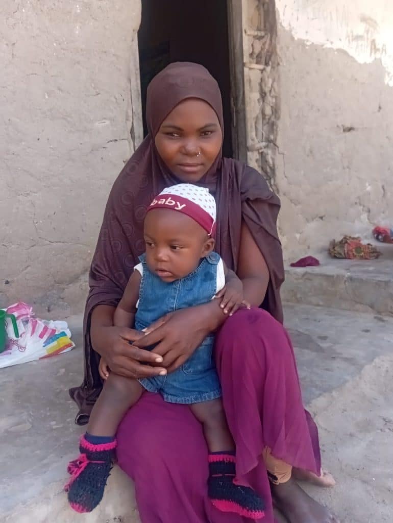 Young African mother sitting outside a simple mud house, holding her baby on her lap, representing motherhood, poverty, resilience, and the daily challenges faced by women raising children in rural communities.