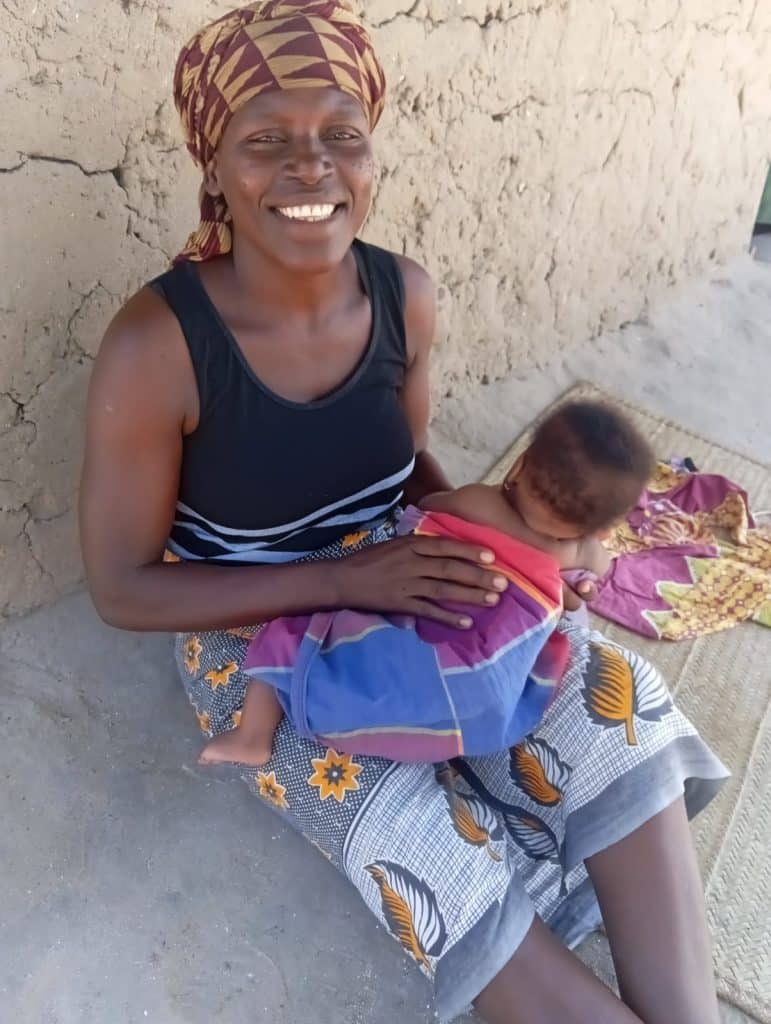frican mother smiling while holding her baby outdoors, sitting on the ground beside a mud wall in a rural community, representing motherhood, resilience, and daily life in a low-income setting.
