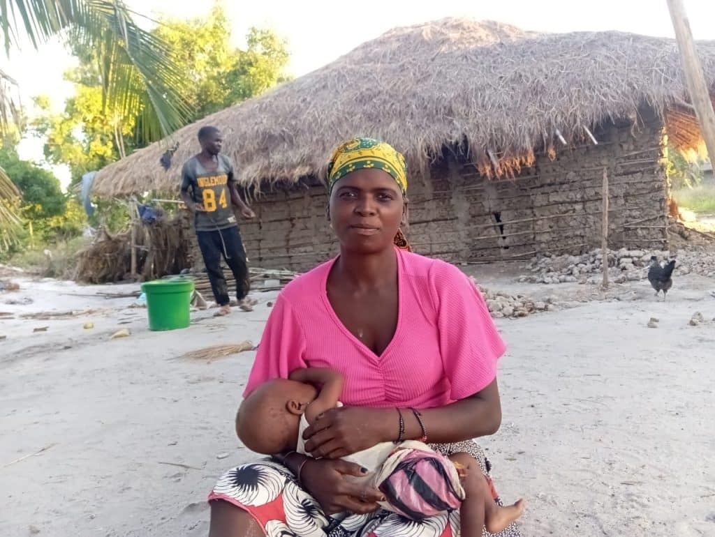 **Alt text:** A young woman sits outdoors in a rural coastal setting, holding and breastfeeding a baby in her arms. She wears a pink top, a patterned skirt, and a green headscarf. Behind her is a traditional mud-and-thatch house, with sandy ground, palm trees, and household items visible, reflecting a simple village environment.